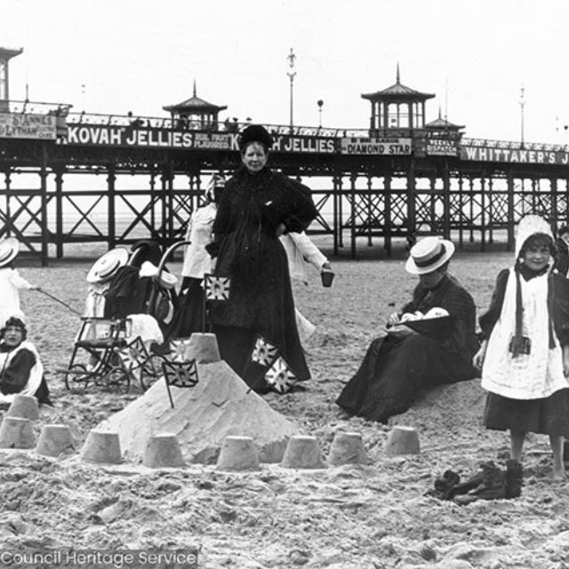 Woman and children building sandcastles, with advertisements on the pier for 'Kovah Jellies, Diamond Star, Weekly Dispatch and Whittakers Jams.'